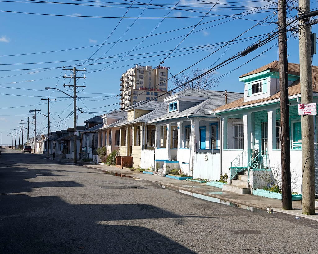 A row of one-story houses painted in pastel. Many powerlines cross in front and a tall apartment building stands behind. 